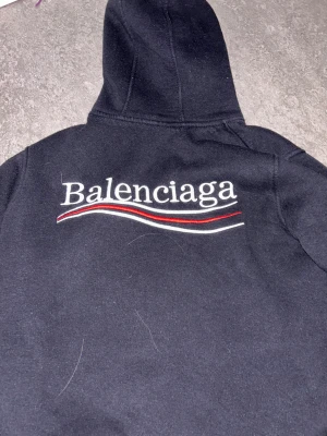 Svart Zara hoddie - Svart hoodie från Zara med broderad logga på bröstet och stor text på ryggen. Klassisk känguruficka framtill, dragsko i huvan och feta röda och vita detaljer i brodyren. Mjuk och skön bomullskänsla, perfekt för en clean streetwear-look.