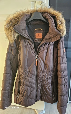 Brun pufferjacka med fuskpäls från Luhta - Säljer en brun pufferjacka från Luhta med snygg fuskpäls på huvan och coola quiltade detaljer. Jackan har dragkedja framtill, två fickor med dragkedja och en klassisk pufferlook som håller dig varm. Perfekt för kalla dagar och riktigt stilren.
