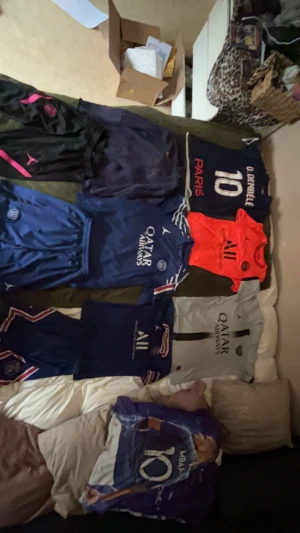 PSG t-shirt med tryck och sponsorlogga - Säljer en PSG t-shirt med tryck av spelare och nummer, samt sponsorlogga på bröstet. Tröjan är kortärmad och finns i flera färger som blå, svart, grå och orange. Perfekt för dig som älskar fotboll och vill representera ditt favoritlag.