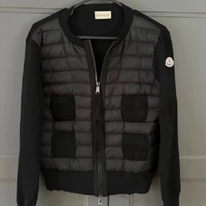 Svart Moncler pufferjacka med stickade ärmar - Snygg svart pufferjacka från Moncler med quiltad front och stickade ärmar. Jackan har dragkedja framtill, rund halsringning och Moncler-logga på ärmen. Materialmix av polyester och stickad ull ger en modern look. Perfekt för dig som vill ha något stilrent och trendigt.