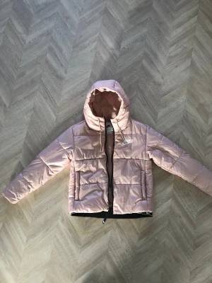 Nike rosa pufferjacka med huva - Säljer en snygg rosa pufferjacka från Nike med Therma-Fit teknologi. Jackan har huva med snörning, dragkedja framtill och två fickor. Perfekt för kyliga dagar och har en modern, boxig passform. Logga på bröstet och quiltad design för extra stilpoäng.