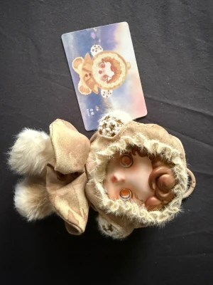 Skullpanda Amber nyckelring Pop Mart - Supersöt Skullpanda Amber nyckelring från Pop Mart Impressionnisme-serien. Nyckelringen har en docka med lockigt brunt hår, fluffig beige huva med pälsdetaljer och stora rosettöron. Materialet är blandat tyg och konstpäls, med gulliga knappar framtill.