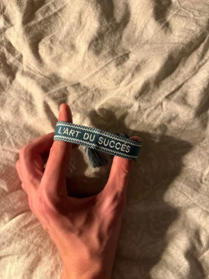 Grönt tygarmband med text - Snyggt grönt tygarmband med vit text 'L'ART DU SUCCÈS' broderat på framsidan. Armbandet har flätade detaljer och tofsar i ändarna för justerbar passform. Perfekt för dig som gillar unika accessoarer med statement.