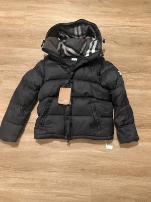 Svart pufferjacka från Burberry M - Svart pufferjacka från Burberry med klassisk rutig huva i grått, vitt och rött. Jackan har dragkedja och knappar framtill, två sidofickor med dragkedja och en snygg Burberry London England-patch på ärmen. Perfekt för kalla dagar med sin fluffiga och quiltade design.