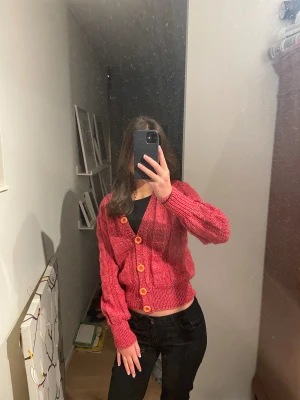 Rosa/röd stickad kofta med knappar - Jättecool stickad kofta med orangea stora knappar. Köpt second hand men bara använt ca 2 gånger själv. Där står ingen storlek eller märke i tröjan men jag skulle uppskatta att den är xs/s. 