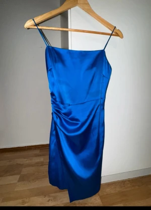 Blå satin klänning från Zara - Snygg blå klänning från Zara i satin med smala axelband och draperad detalj på sidan. Modellen är kort och har en stilren, figurnära passform som ger en elegant siluett. Perfekt för dig som vill sticka ut med en färgstark look.väldigt skön material.
