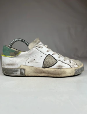 Philippe model vita Stl 40 - Säljer ett par vita Philippe Model storlek 40 sneakers med grå mockadetaljer och snörning. Skorna har en rund tå, platt sula och en unik patch på sidan. Bakkappan är grön med camomönster och gul kant. Perfekt för dig som gillar streetstyle och exklusiva märken. Hör gärna av dig vid frågor eller funderingar. 