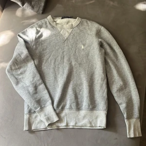 Grå sweatshirt från Polo Ralph Lauren - Klassisk grå sweatshirt från Polo Ralph Lauren med broderad logga på bröstet. Tröjan har rund halsringning, ribbade muddar och är tillverkad i mjuk bomullsmix. Perfekt för en chill och stilren look.