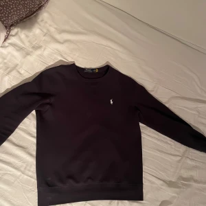 Svart tröja från Polo Ralph Lauren - Klassisk mörkblå tröja från Polo Ralph Lauren med rund halsringning och broderad vit logga på bröstet. Tröjan har lång ärm och är tillverkad i mjuk bomull, perfekt för en stilren och enkel look.
