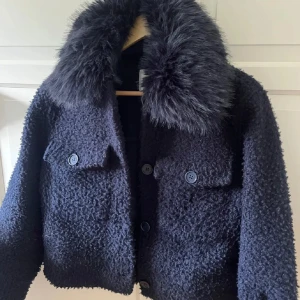 Mörkblå teddyjacka med päls - Säljer en mörkblå teddyjacka med fluffig päls på kragen. Jackan har två stora fickor med knappar framtill och en croppad passform. Perfekt för dig som vill ha en cool och mysig look under kyliga dagar.