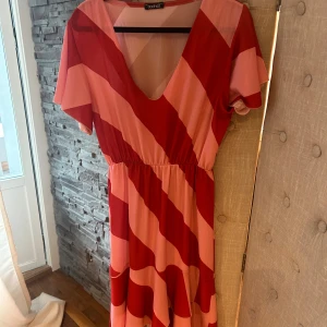 Randig röd och rosa klänning Boohoo - Säljer en långklänning från Boohoo med breda diagonala ränder i rött och rosa. Klänningen har v-ringning, korta ärmar och markerad midja med resår. Luftigt material, perfekt för varma dagar. Snygg och färgstark statement-piece!