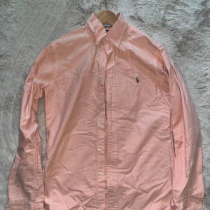Rosa skjorta från Ralph Lauren - Snygg rosa skjorta från Ralph Lauren med klassisk button-down krage och broderad logga på bröstet. Skjortan har lång ärm, knappar framtill och är gjord i mjuk bomull. Perfekt för dig som gillar stilrena och fräscha plagg med en modern passform.