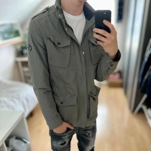 Grå vindjacka med fickor - Snygg grå vindjacka med flera stora fickor framtill, axelklaffar och hög krage. Jackan har knappar och dragkedja samt en cool patch på ärmen. Perfekt för dig som gillar en stilren och funktionell look.