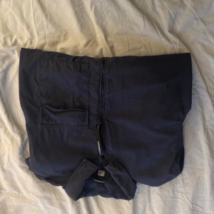 Svart overshirt/vindjacka från C.P. Company - Säljer en jävligt snygg svart vindjacka/overshirt från C.P. Company med dragkedja framtill, klassisk krage och en ficka på bröstet. Jackan har lång ärm med den ikoniska linsen på ena ärmen. Tillverkad i polyester och har en clean, stilren look. Ett av mitt bästa köp