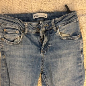 LOW WAIST Blå bootcut jeans från Zara - LOWWAIST Snygga ljusblå jeans från Zara med klassisk femficksdesign och bootcut-ben. Jeansen har slitningar på ena benet och bakfickan, vilket ger en cool och avslappnad vibe. Tillverkade i mjukt denim-material med dragkedja och knapp framtill. Tveka inte på att höra av dig vid frågor eller intresse!