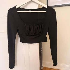 Svart croppad topp  - Svart långärmad croppad topp från SHEIN med fyrkantig ringning och rynkade detaljer framtill. Tunn och stretchig i materialet, perfekt att styla med höga jeans eller kjol. Enkel men trendig design som funkar till många looks.