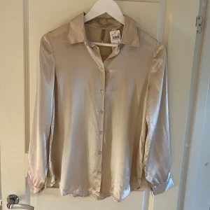 Champagnefärgad silkesblus Intimissimi - Snygg champagnefärgad skjorta från Intimissimi i lyxigt silke. Klassisk krage, långärmad och med knappar framtill. Perfekt för dig som gillar en clean och elegant look. Skjortan har en mjuk glans och känns superlätt mot huden. Helt ny med prislappen kvar köpt för 89,90 euro som är 980kr 