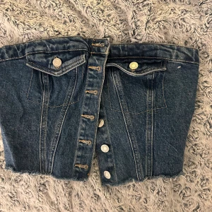 Jeans bandeau tubtopp med knappar - Säljer en cool jeans bandeau tubtopp med knappar framtill, fickdetaljer och rå kant nertill. Baksidan har smock för extra stretch och passform. Perfekt för dig som vill sticka ut med en edgy denimlook.