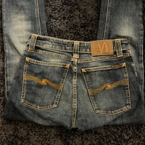 Blå jeans från Nudie - Säljer ett par klassiska blå jeans från Nudie Jeans med tydliga slitningar och snygga gula sömmar på bakfickorna. Jeansen har fem fickor, normal passform och är tillverkade i denim. Storlek är 27/32 med en normal passform som varken är för lös eller tajt.