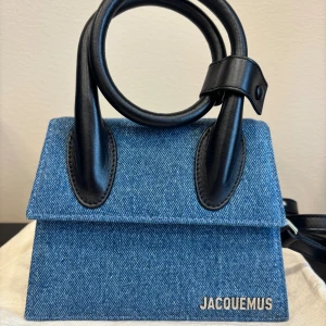 Blå denimväska från Jacquemus - Superfin liten handväska från Jacquemus i blått jeanstyg med svarta detaljer i skinn. Väskan har ett stilrent, fyrkantigt format och ett runt handtag i svart skinn. Snygg Jacquemus-logga i metall framtill och magnetlås. Perfekt för dig som gillar trendiga accessoarer. Aldrig använd. Dustbag och etikett medföljer. 