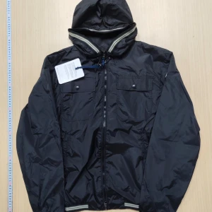 Svart vindjacka från Moncler med huva - Svart vindjacka från Moncler med huva, dragkedja framtill och två bröstfickor med knappar. Jackan har ribbade muddar med ränder och Moncler-logga på ärmen. Tillverkad i lätt och glansig polyester, perfekt för blåsiga dagar. Kvitto och påse medföljer. Jackan är aldrig använd.
