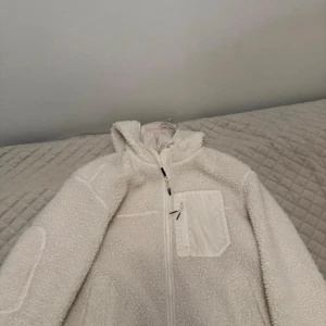 Vit teddyjacka med huva - Mysig vit teddyjacka med huva och dragkedja framtill. Jackan har en praktisk bröstficka med dragkedja och är tillverkad i fluffigt teddy-material som håller dig varm. Perfekt för kyliga dagar och enkel att styla till olika outfits.