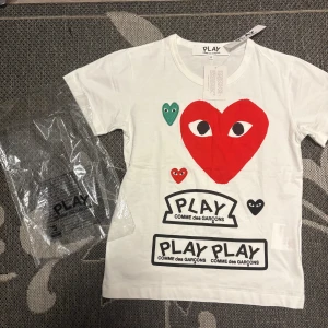 Vit Comme des Garçons Play t-shirt - Vit t-shirt från Comme des Garçons Play med stora och små hjärtan i rött, grönt och svart framtill. Ikonisk logga och tryck, samt broderade detaljer. Klassisk passform och rund hals. Tillverkad i 100% bomull för en skön känsla.
