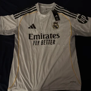 Real Madrid vit matchtröja Adidas - Säljer en officiell Real Madrid fotbollströja från Adidas i vitt med svarta detaljer och gula linjer. Tröjan har korta ärmar, klubbmärke på bröstet och sponsortryck. Tillverkad i lätt och ventilerande material, perfekt för match eller träning. Storlek S men passar M också.