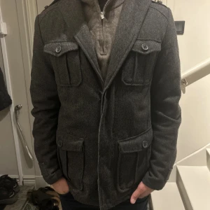 Mörkgrå ullrock med fickor - Stilren mörkgrå rock i ull med fyra stora fickor framtill och snygga knapplock. Klassisk krage och långärmad modell, perfekt för kyliga dagar. Snygg och tidlös design som passar till det mesta.