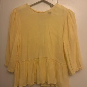 Gul volangblus med knyt i ryggen - Söt ljusgul blus med volangkant och trekvartsärmar. Baksidan har två knytband som ger en öppen rygg-detalj. Blusen är i ett lätt, krinklat bomullstyg och har en luftig, loose passform. Perfekt för vår och sommar!