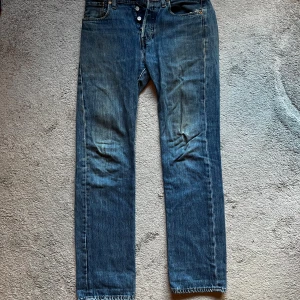 Levi's 501 blå jeans straight fit - Klassiska Levi's 501 jeans i blå tvätt med raka ben och knappgylf. Jeansen har fem fickor, brunt läderpatch bak och snygga slitningar för en cool vintagekänsla. Perfekt för dig som gillar tidlös streetstil och vill ha ett par jeans som funkar till allt. Skriv för mer bilder