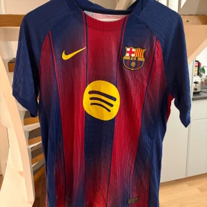 FC Barcelona Lamine Yamal matchtröja - Säljer en officiell FC Barcelona fotbollströja från Nike med Lamine Yamal och nummer 10 på ryggen. Tröjan har klassiska blå och röda vertikala ränder, gula detaljer och klubbmärke på bröstet. Tillverkad i lätt och ventilerande polyester, perfekt för match eller träning.