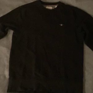 Svart sweatshirt från GANT, storlek M - Svart sweatshirt från GANT i klassisk modell med rund halsringning och diskret broderad logga på bröstet. Tröjan har långa ärmar och ribbade muddar vid ärmslut och nederkant. Den är i bra skick för attJag har inte använt den knappt för jag inte tyckte att de passade på mig så den är bara använd 2-3 gånger. (Pris kan diskuteras)
