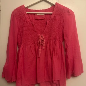 Rosa blus med knyt och smock - Supersöt rosa blus från Odd Molly med smockdetaljer över bröst och rygg, v-ringning med knytband och vida långa ärmar. Blusen har en lätt volang nertill och är gjord i mjuk bomull med diskret prickstruktur. Perfekt för dig som gillar boho-vibbar!