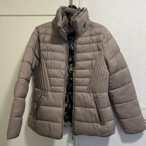 Beige pufferjacka från Flash Woman - Säljer en beige pufferjacka från Flash Woman i storlek S. Jackan har quiltade detaljer, hög krage och dragkedjor på fickorna. Perfekt för kyliga dagar med sin vadderade design och stilrena look.