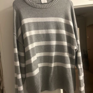 Gråvit randig stickad tröja - Mysig stickad tröja i grått och vitt med breda horisontella ränder. Tröjan har rund halsringning med ribbad kant och långa ärmar. Perfekt för lager-på-lager och chill dagar. Oversized fit för en avslappnad look.
