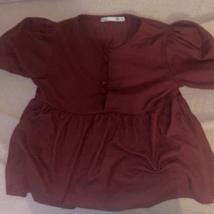 Vinröd blus med puffärm från 157 - Supersöt vinröd blus från 157 med puffiga korta ärmar och två knyt framtill. Blusen har en lätt rynkad nederdel och är gjord i ett mjukt, strukturerat tyg. Perfekt för dig som vill ha en chill men ändå stylish look.