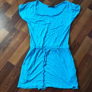 Turkos kort klänning från Warp - Turkos kortärmad klänning från Warp i mjuk bomull. Modellen är kort med rund halsringning och har ett flätat knytband i midjan för en snygg passform. Perfekt för varma dagar och enkel att styla med sneakers eller sandaler.