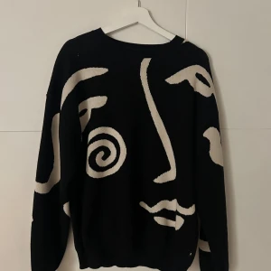 Svart stickad tröja med ansiktsmönster - Cool svart stickad tröja från Sweet SKTBS med ett stort, grafiskt ansiktsmönster i beige på framsidan. Tröjan har rund hals och långa ärmar, perfekt för dig som gillar unik design och streetstyle. Ribbstickade muddar vid hals och ärmslut.
