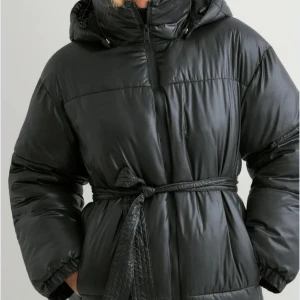 Svart pufferjacka från Nelly med huva - Svart pufferjacka från Nelly i storlek S. Jackan har en stor, mysig huva, dragkedja framtill och ett snyggt bälte i midjan. Materialet är glansigt och quiltat, perfekt för kalla dagar. Jackan har en loose passform och långa ärmar.Jackan är i nyskick, endast använd 2–3 gånger. Inköpt i vintras