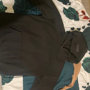 Svart Essentials hoodie med huva - Svart hoodie från Essentials Fear of God med stor logga på ryggen och mindre tryck framtill. Klassisk känguruficka och huva. Perfekt för dig som gillar streetwear och vill ha en stilren look.