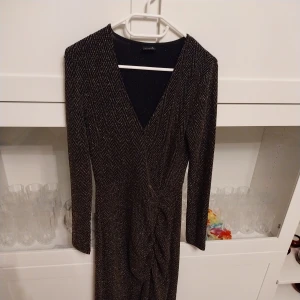 Glittrig svart långklänning med v-ringning från Gina Tricot  - Snygg svart långklänning med glittriga detaljer och v-ringad hals. Klänningen har lång ärm, omlott-effekt framtill och ett subtilt mönster som ger extra stilpoäng. Perfekt för dig som vill sticka ut med en elegant look. Storlek:M (Medium)