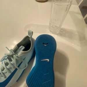 Nike blåvita fotbollsskor - Säljer ett par snygga Nike fotbollsskor i ljusblått och vitt med blå sula och klassisk Nike-logga på sidan. Skorna har snörning och är designade för att ge bra grepp på planen. Perfekta för dig som vill sticka ut med stilrena och sportiga skor.