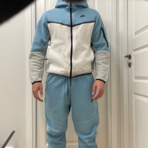 Nike ljusblå tracksuit M och L - Nike mjukisbyxor och hoodie i ljusblå färg med svart logga på vänster lår. Byxorna har normal passform passar både L och M elastisk midja med snörning och ribbade muddar vid benslut. Hoodie i storlek M. Tillverkade i mjukt bomullsmaterial som är skönt mot huden. Perfekta för chill eller träning. 