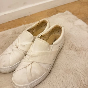 Vita slip-on sneakers med knutdetalj - Snygga vita slip-on sneakers från Anna Field med unik knutdetalj framtill. Skorna har en rund tå, platt sula och är tillverkade i textil. Insidan är dekorerad med små svarta prickar för en extra touch. Perfekta för en clean och stilren look.