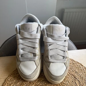 Vita Adidas originals sneakers i mocka och skinn - Säljer ett par stilrena Adidas sneakers i vitt med detaljer i ljusgrå mocka och vitt skinn. Skorna har breda vita snören, rund tå och klassisk Adidas-logga på plösen och sidan. Perfekta för dig som gillar en clean och sportig look.
