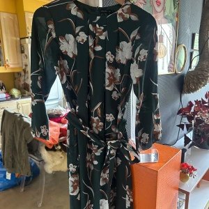 Blommig klänning från STOCKHLM - Mörkgrön knälång klänning med stora blommor i vitt och brunt från Stockhlm. Klänningen har trekvartsärm, knytband i midjan och rund halsringning med veckdetaljer. Använd 1 gång. 