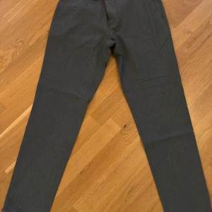 Mörkgrå kostymbyxor från Bläck - Snygga mörkgrå kostymbyxor från Bläck i modellen Tailored Fit. Byxorna har en klassisk rak passform och är perfekta för en stilren look. Tillverkade i ett mjukt material som känns bekvämt hela dagen. Passar dig som gillar en dressad och modern stil.