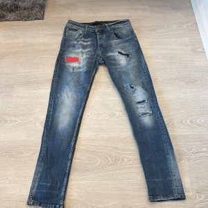 Philipp Plein blå slitna jeans, stl 30 - Blå jeans från Philipp Plein med coola slitningar, trasiga detaljer och en röd patch på låret. Jeansen har klassisk femficksdesign, normal passform och är tillverkade i denim. Snyggt tvättade med ljusare partier och unika detaljer som sticker ut.