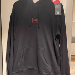 Svart hoodie från Hugo med röd logga - Svart hoodie från Hugo med röd logotyp på bröstet. Tröjan har huva med snörning, stor magficka och långa ärmar. Materialet är mjuk bomull som känns skönt mot huden. Perfekt för dig som gillar stilren design med en touch av färg.